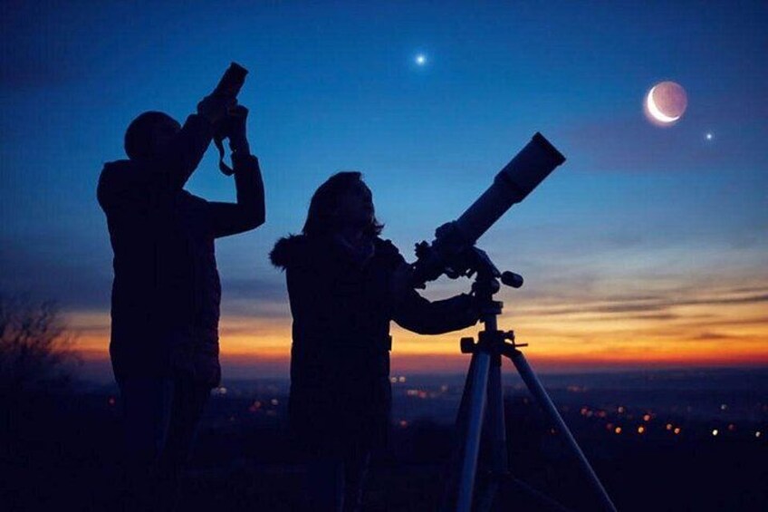 Dos personas usando telescopios de noche, una luna creciente y un cielo estrellado. 1