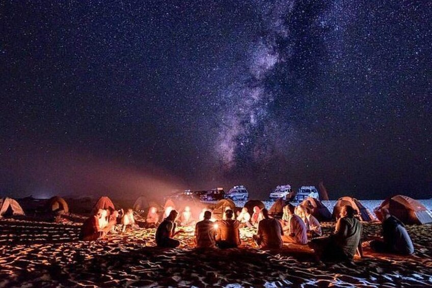 Cielo nocturno estrellado con la Vía Láctea visible, personas sentadas alrededor de una fogata y tiendas de campaña instaladas en una playa. 5