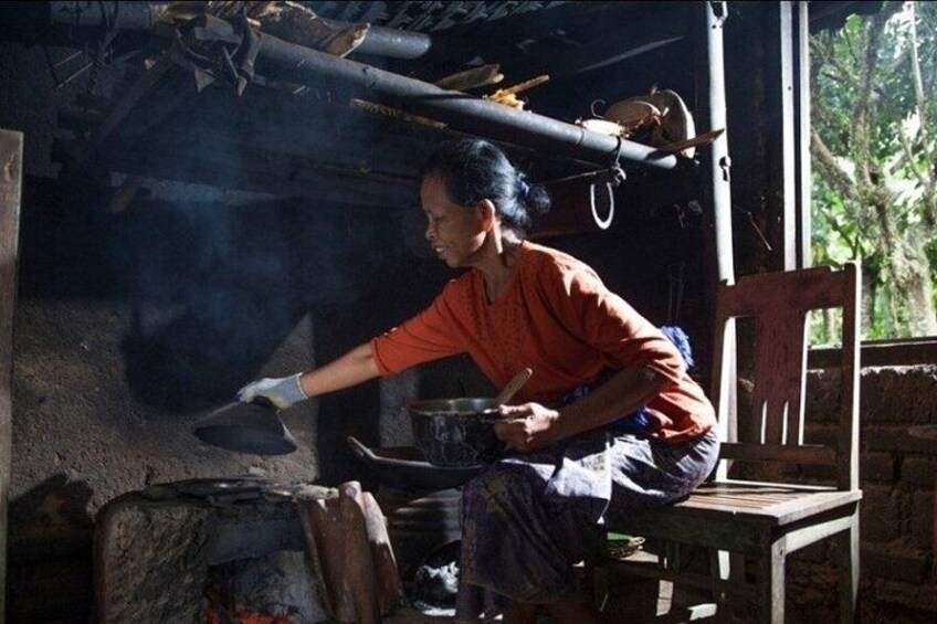 A person cooking in a rustic kitchen with a large pot on a stove, a wooden chair, and a brick wall. 3
