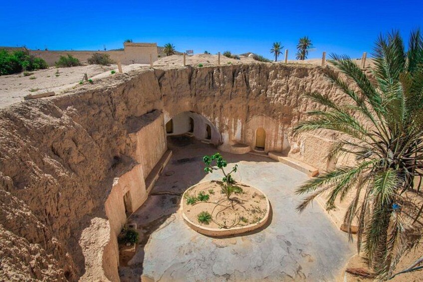 An ancient archaeological site with a large open courtyard, a palm tree, and a small building. 3