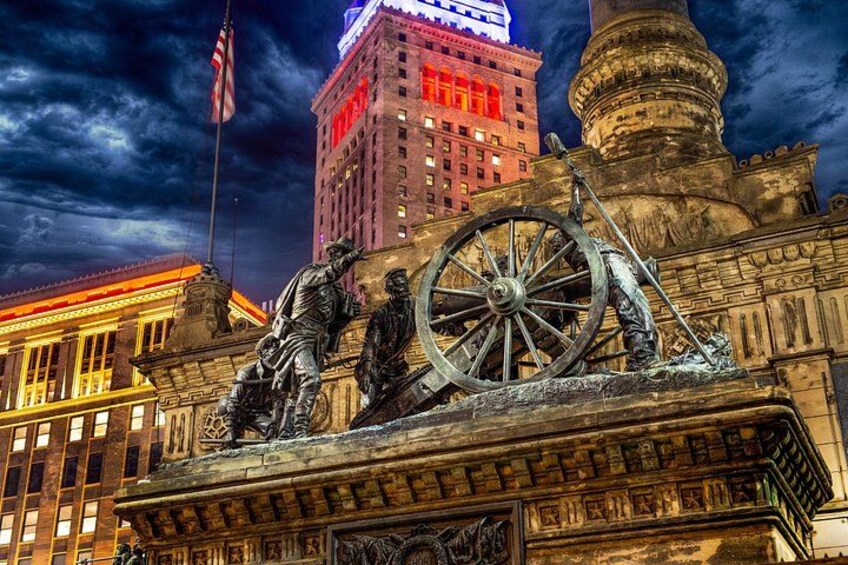 A historical monument with a large wheel and statues, a tall building with illuminated windows, and an American flag. 2
