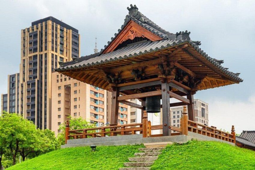 Een traditioneel Koreaans paviljoen met pannendak, omgeven door een pad van stenen en weelderige begroeiing, met op de achtergrond moderne wolkenkrabbers. 2