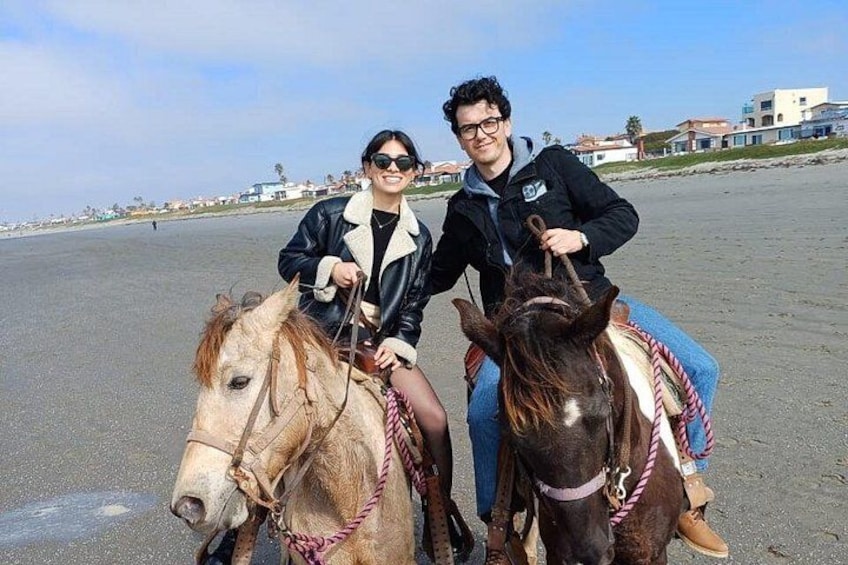 Two people on horseback on a beach with houses in the background. 3