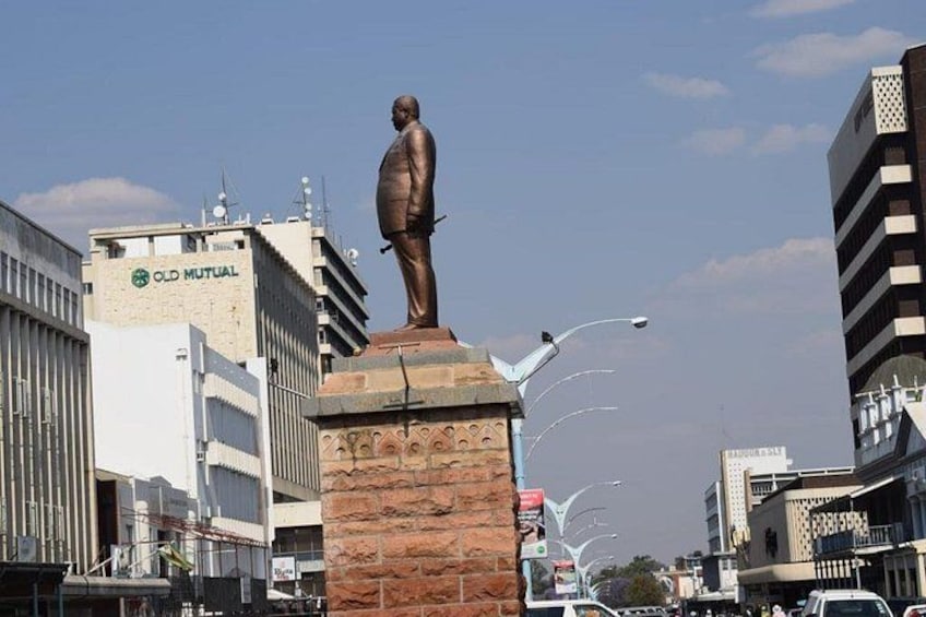 A statue on a pedestal in an urban setting with buildings and a clear sky. 1