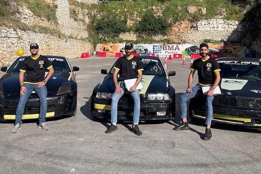 Three men sitting on race cars with 'DRIFT KING' branding, parked on a paved surface with a stone wall and trees in the background. 2