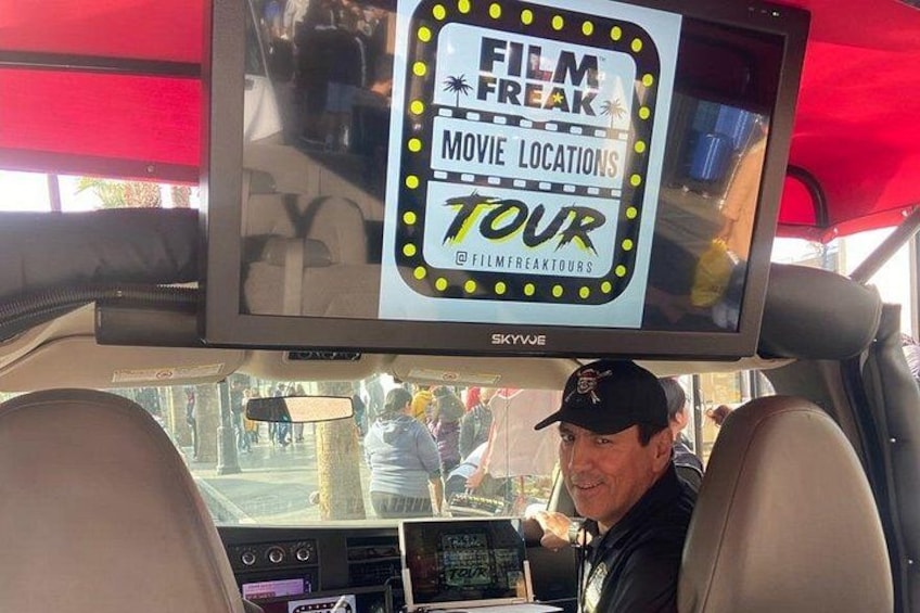 A vehicle interior with a screen displaying 'FILM FREAK MOVIE LOCATIONS TOUR' and a person wearing a cap. 2