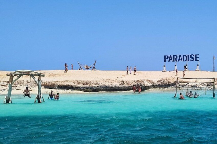 A beach with clear turquoise water, a swing set, and the word 'PARADISE' on the sand. 4