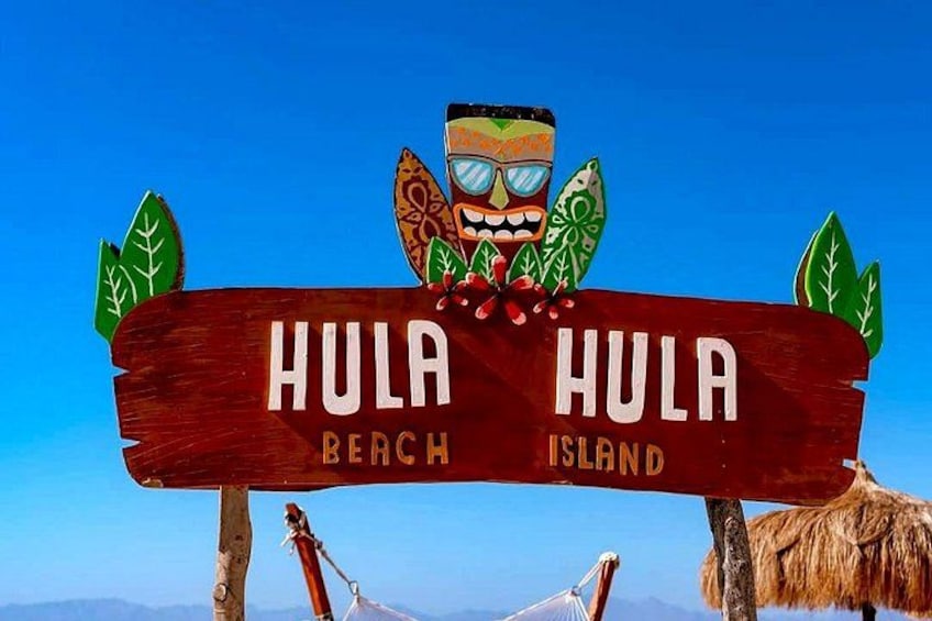 A wooden sign with 'Hula Beach Island' written on it, a tiki head sculpture, and a thatched hut. 1