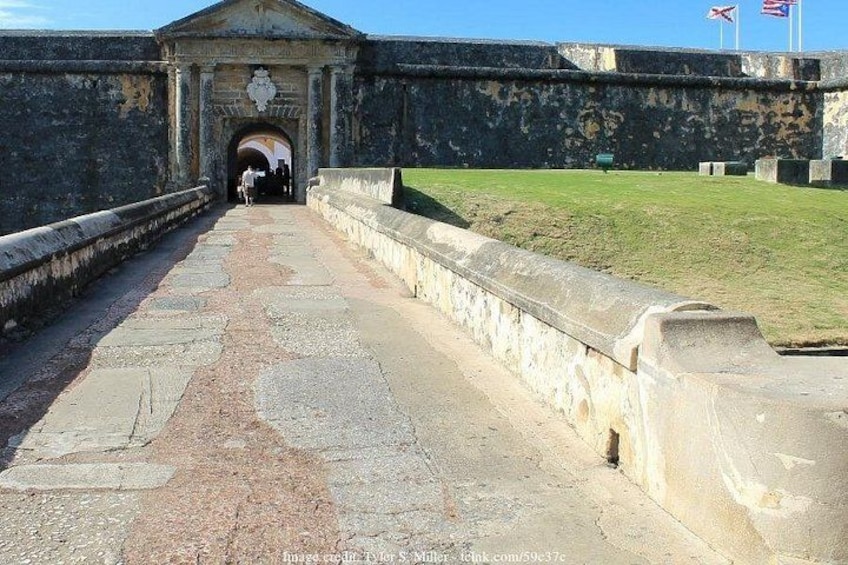 A historic fort entrance with a paved pathway leading to a gate, surrounded by stone walls and a grassy area. 2
