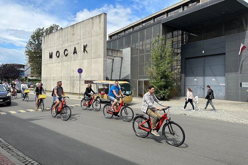 En gade med cyklister, en bygning med teksten 'MOCAK' og et fodgængerfelt. 1