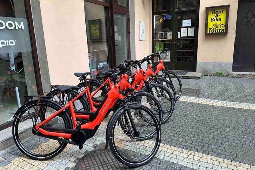 Række af røde elektriske cykler parkeret foran en bygning med et skilt til cykelture. 4