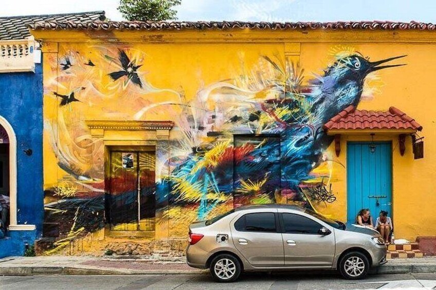 A building with a vibrant mural of a bird and abstract patterns, a blue door, and a parked car. 5