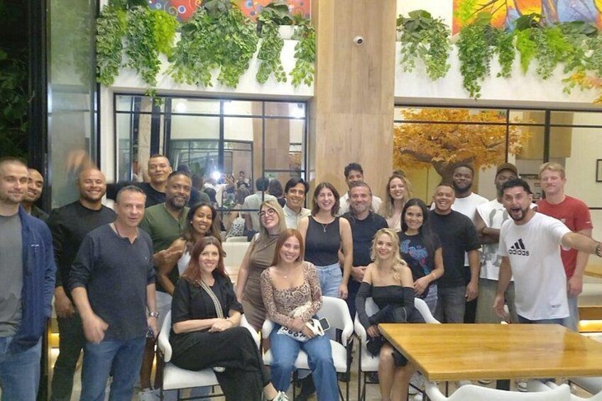 A group of people posing indoors with tables and chairs, surrounded by greenery and a large window. 4