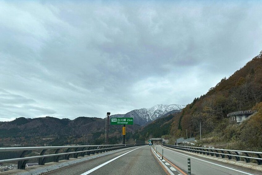 A highway with guardrails, a green road sign, and snow-capped mountains in the background. 4