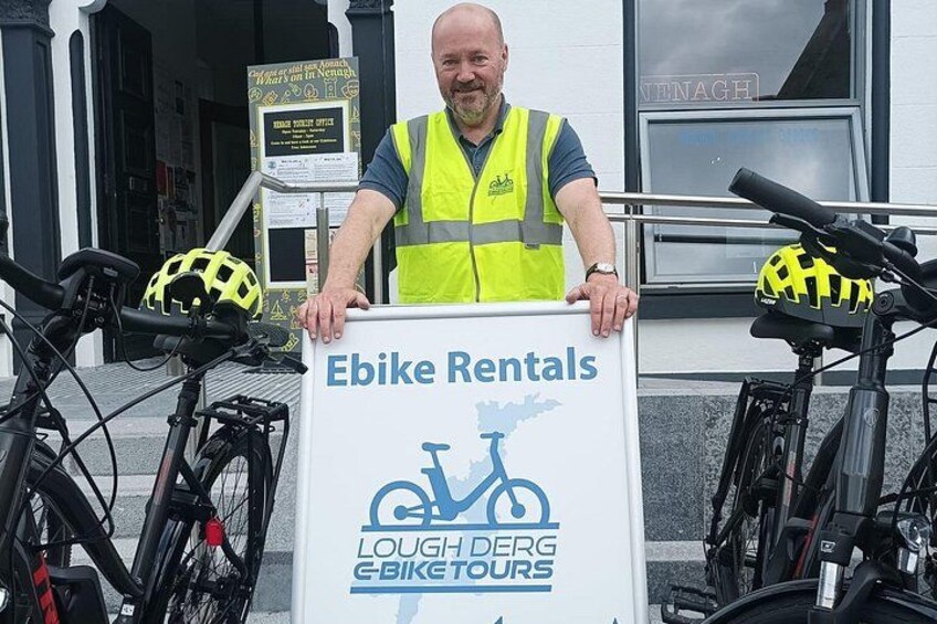 Een man houdt een bord vast voor Ebike Verhuur voor een gebouw met fietsen. 5