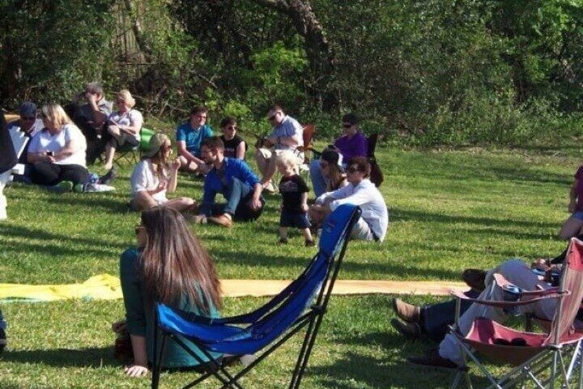 People sitting on grass in a park, some on chairs, others on the ground, with trees in the background. 3