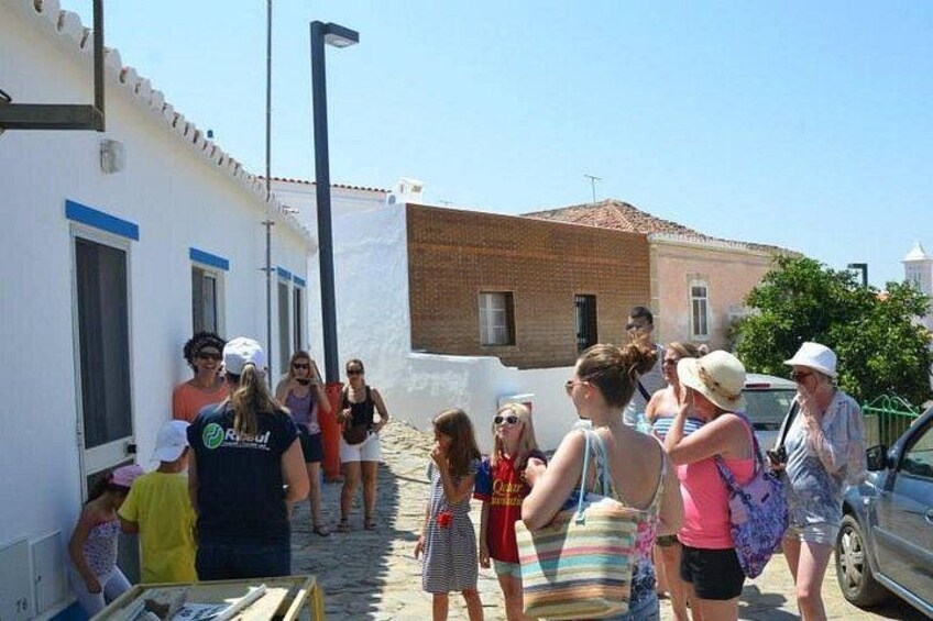 Un groupe de personnes se tient dans une rue, certaines avec des sacs, près d’un bâtiment blanc orné de motifs bleus, et un bâtiment en briques se trouve à l’arrière-plan. 3