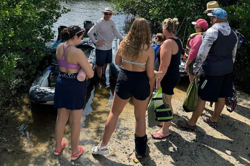 A group of people standing by a river, some holding bags, with a boat in the background. 5