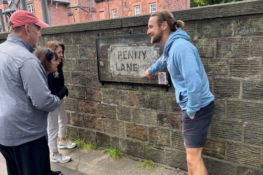 A group of people standing by a stone wall with a sign that reads 'PENNY LANE'. 2