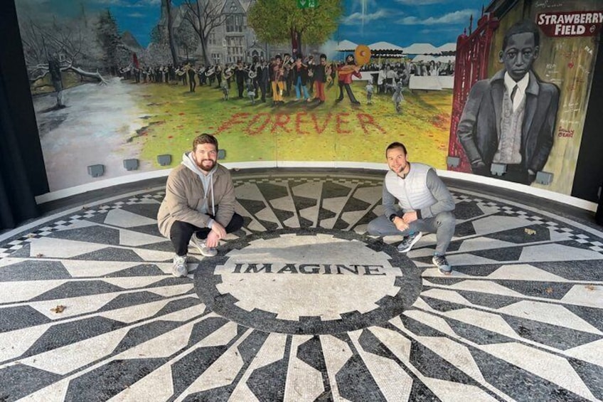 Two people sitting on a patterned floor with the word 'IMAGINE' in the center, surrounded by a mural with a large tree and a building in the background. 1