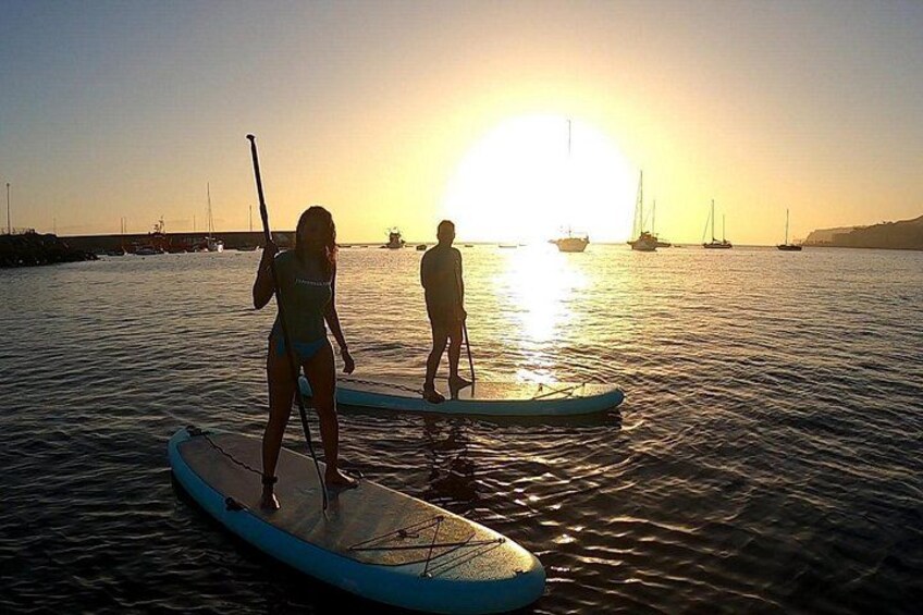 To personer paddleboarder på roligt vand i solnedgangen, med både i baggrunden. 4
