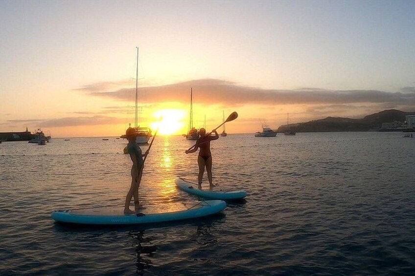 To personer paddleboarder på roligt vand i solnedgangen, med både i baggrunden. 3