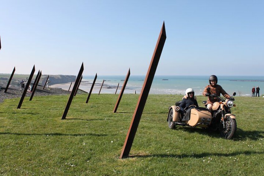 Eine grüne Wiese mit Metallpfählen, einem Motorrad aus vergangenen Zeiten mit Beiwagen und einem Strand im Hintergrund. 2