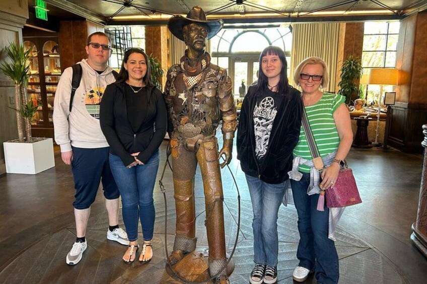 Un grupo de personas posando con una estatua de bronce en un vestíbulo con pisos de madera y ventanas amplias. 3