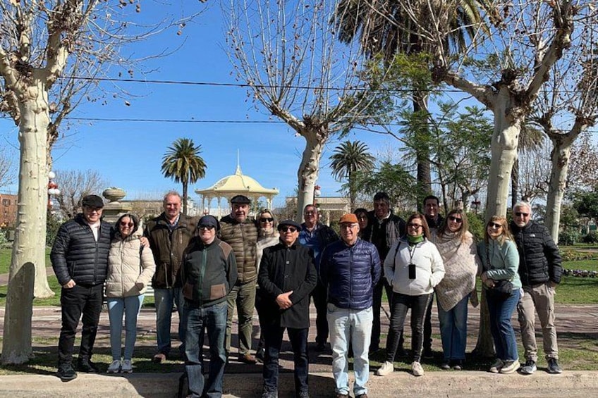 A group of people standing in a park with trees and a gazebo in the background. 2