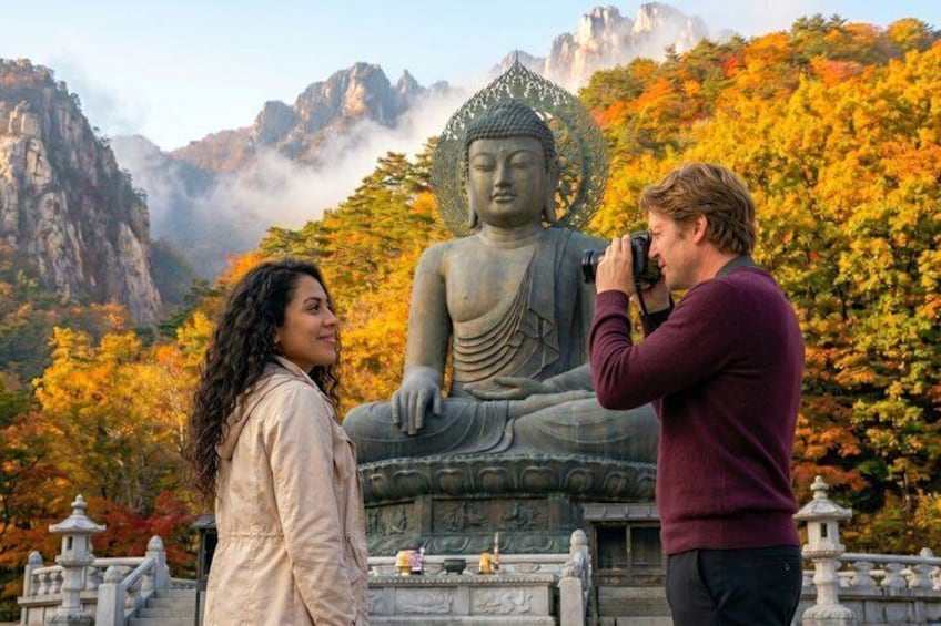 A person taking a photo of a large Buddha statue with autumn trees in the background. 1