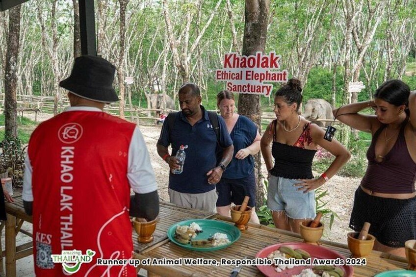 A group of people at Khao Lak Ethical Elephant Sanctuary, with a man in a red vest labeled 'Khao Lak Thailand' and a sign for the sanctuary. 5