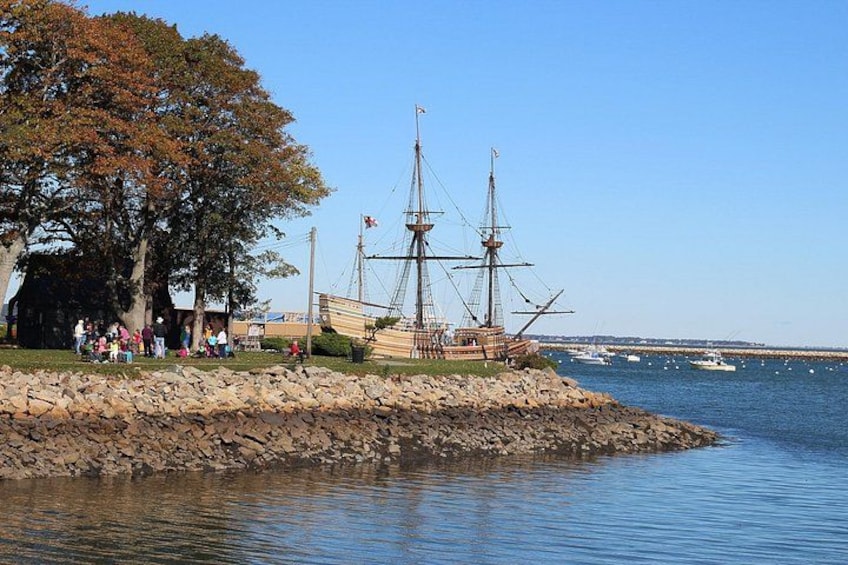 A historic ship docked at a waterfront with people gathered around, a rocky shoreline, and calm water. 4