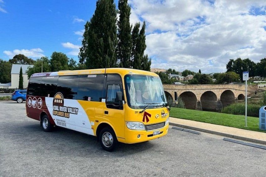 Een gele bus met daarop 'Coal River Valley Explorer Bus' geparkeerd aan de kant van de weg, met een brug op de achtergrond. 2