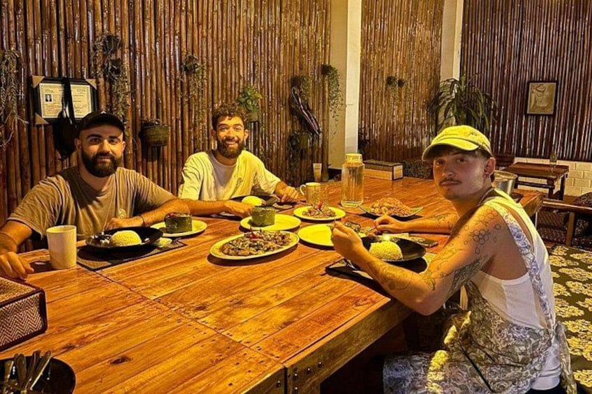 Three people sitting at a wooden table with plates of food, in a room with bamboo walls. 4