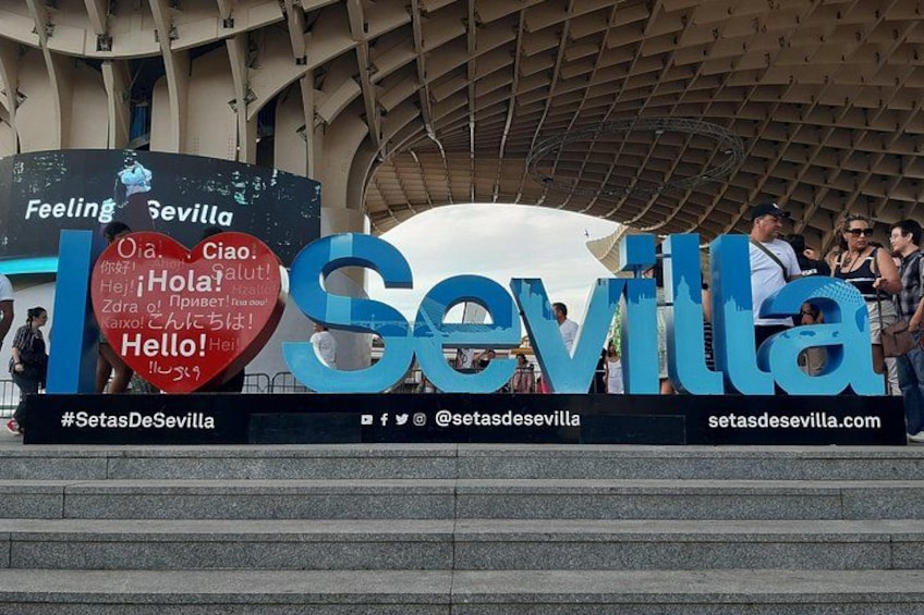 A large outdoor sign with 'Sevilla' written in blue letters, a heart-shaped sign with greetings in multiple languages, and a hashtag #SetasDeSevilla. 4