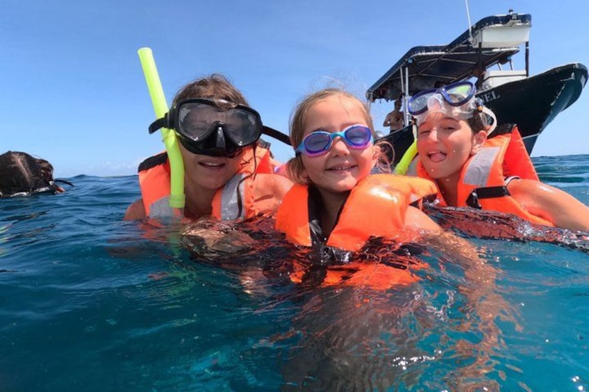 Tres personas haciendo snorkel en aguas azules y cristalinas, con un barco al fondo. 3