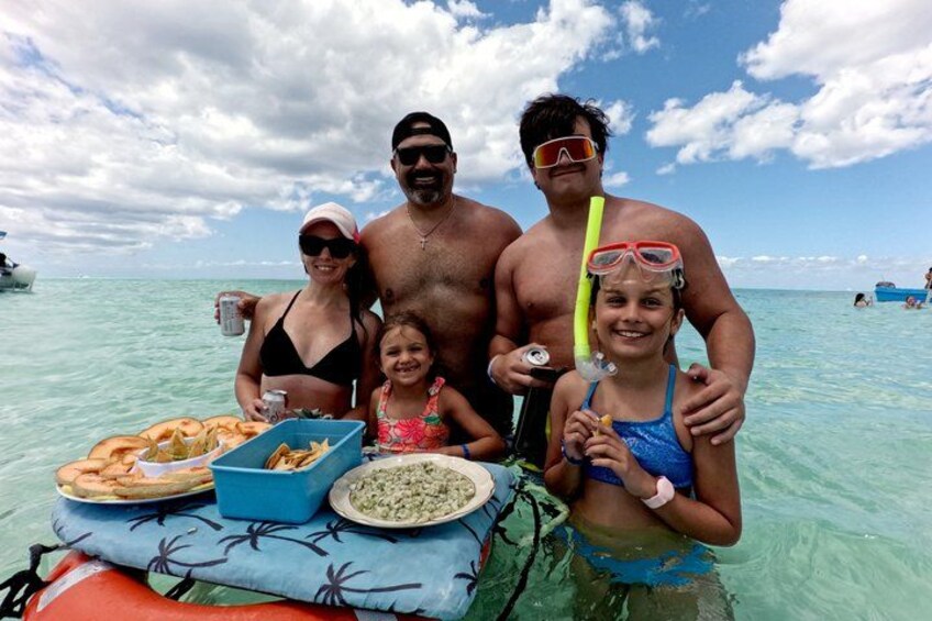 Una familia en un bote, con refrigerios y bebidas, rodeada de agua turquesa cristalina y un cielo parcialmente nublado. 4