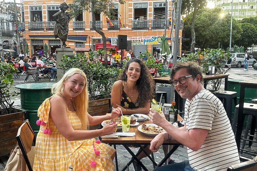 Tre personer spiser på en udendørs café med en statue og bygninger i baggrunden. 1