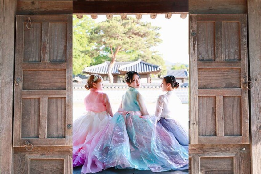 Three children in colorful dresses sitting on a wooden bench in front of a traditional Korean gate. 1