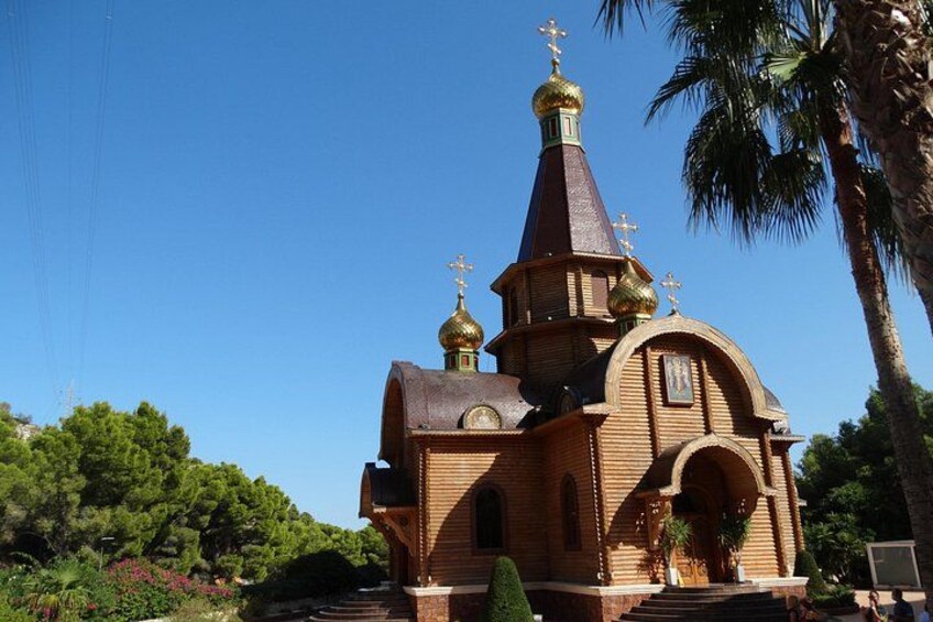 En kyrka byggd av trä, med ett högt klocktorn och kors på toppen, omgiven av grönska. 3