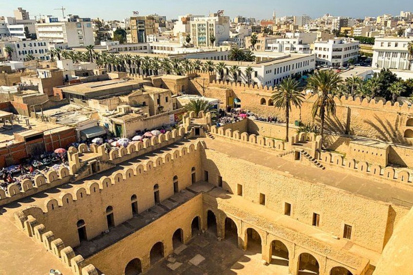 A historical fortress with arched walkways and a surrounding wall, overlooking a modern cityscape with palm trees and buildings. 2