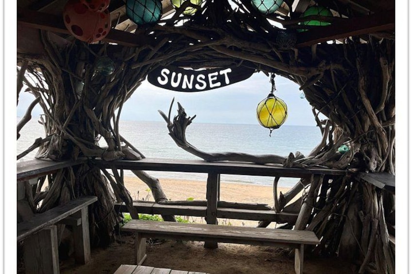 A wooden bench under a rustic structure with a 'SUNSET' sign, overlooking a beach. 5
