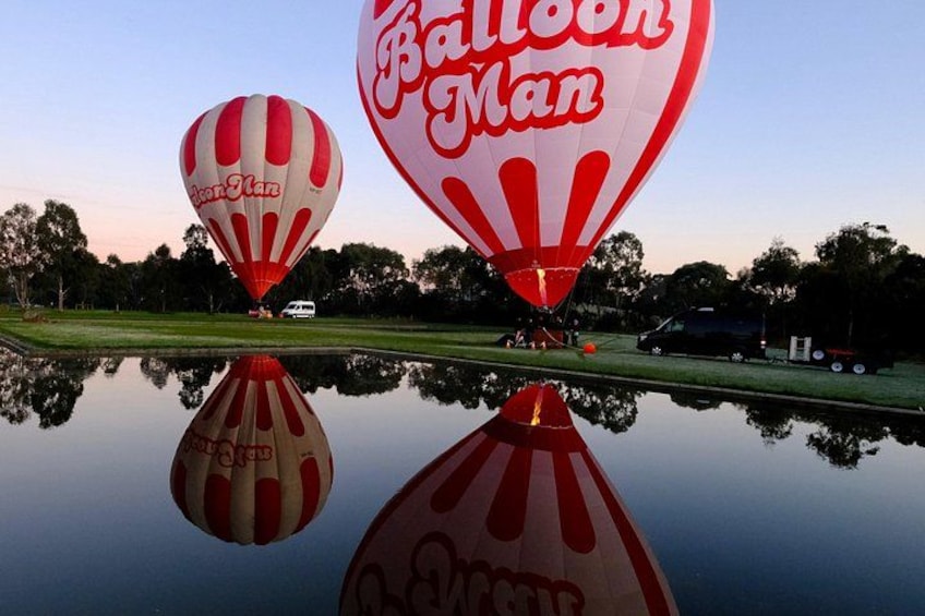 Two hot air balloons with 'Balloon Man' branding over a calm water body with reflections, surrounded by trees and a clear sky. 4