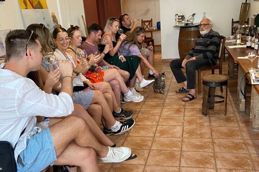 A group of people seated in a room with a tiled floor, a wooden table with bottles, and a cat on the floor. 4