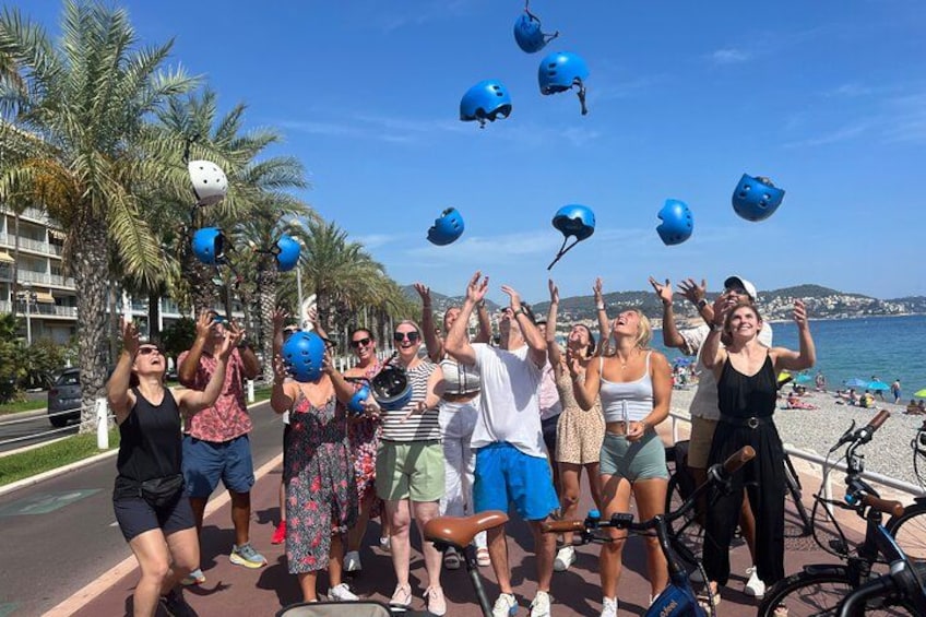 Eine Gruppe Menschen am Strand, einige tragen Helme, im Hintergrund Fahrräder und Palmen. 1