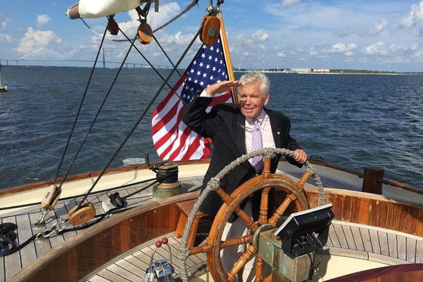 A person steering a wooden boat with an American flag, a steering wheel, and a view of a bridge in the background. 1