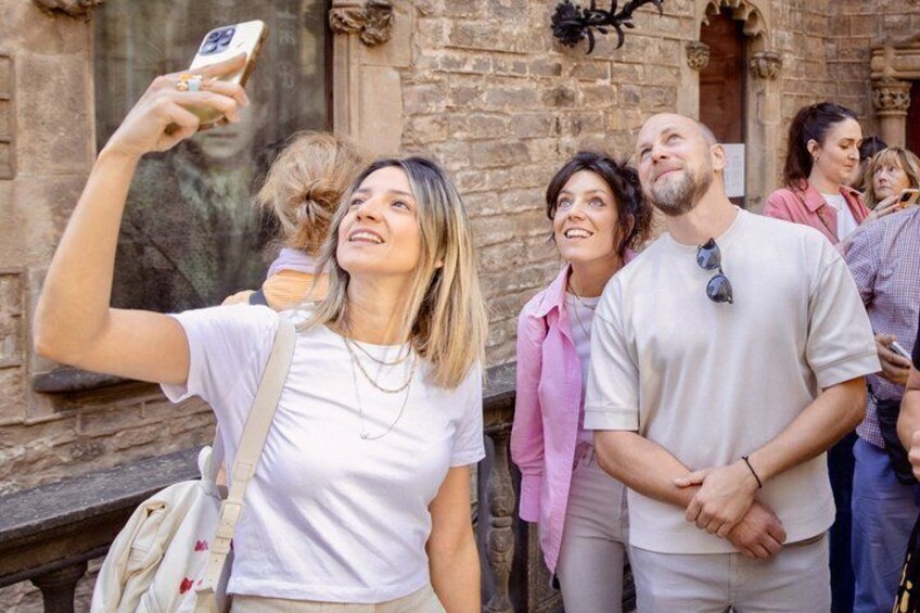 A person taking a selfie with a child on their back, a stone building in the background, and a group of people standing nearby. 2