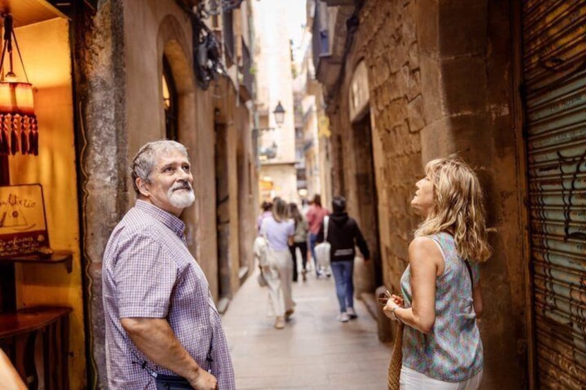 A narrow street with buildings on either side, a man and a woman standing and conversing, and people walking in the background. 4
