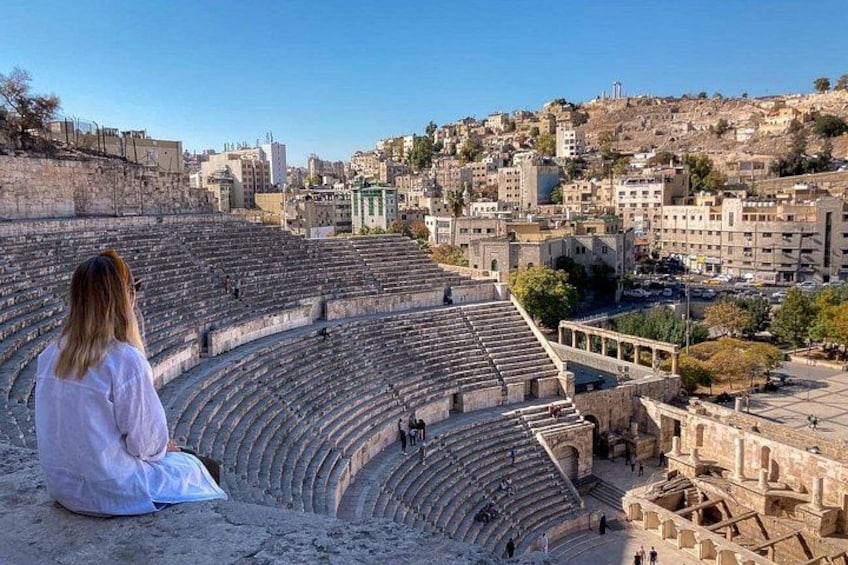 Un anfiteatro antiguo con asientos de piedra, una persona sentada en los escalones y un paisaje urbano al fondo. 1