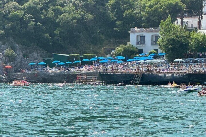 Una playa concurrida donde gente nada y toma el sol, sombrillas azules y una costa rocosa. 4